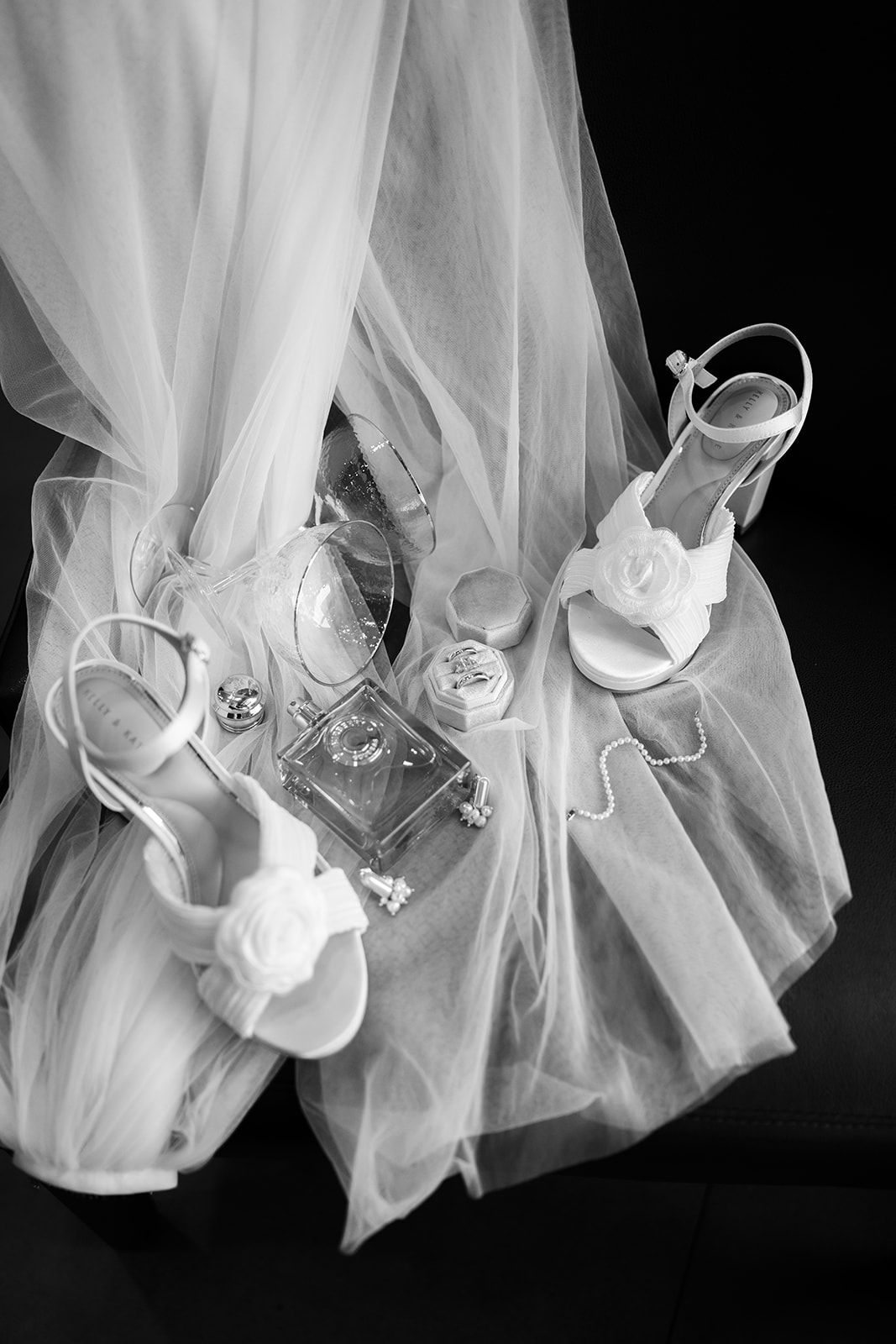 Black and white photo of bridal details, including white floral heels and perfume, arranged on a soft veil.